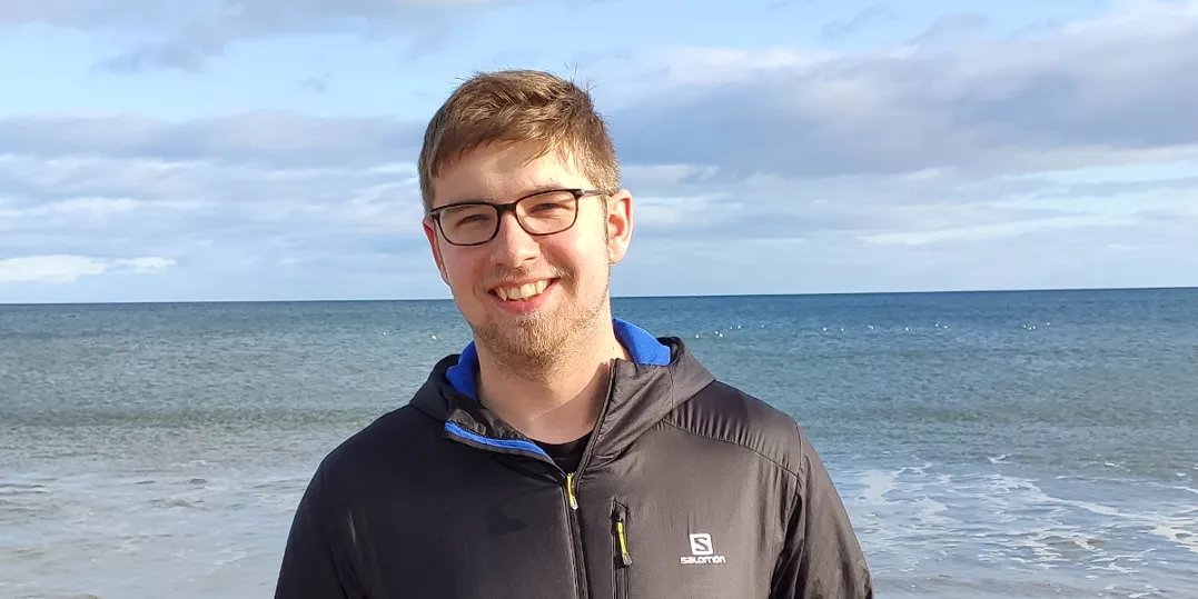 Photo of me smiling at the camera in front of a background of a slightly rough sea and cloudy sky.