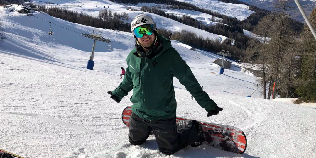 Photo of me kneeling on a snowy ski slope, with a snowboard on my feet. I'm smiling at the camera in full ski kit, my arms are outstretched with two thumbs up.
